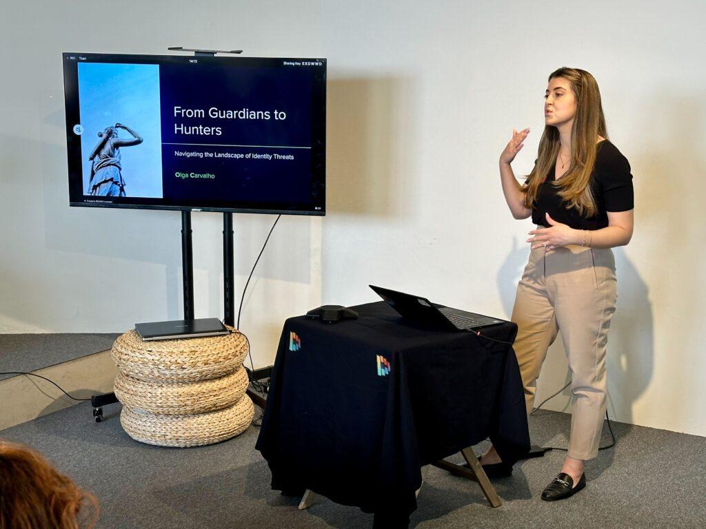 Olga Carvalho standing to present in front of a monitor that reads “From Guardians to Hunters: Navigating the Landscape of Identity Threats.