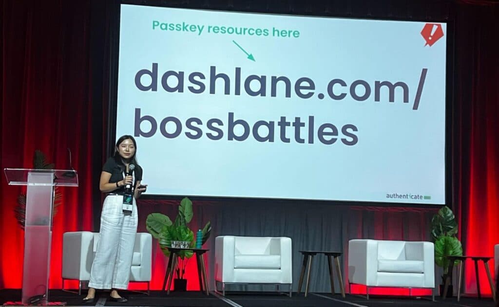 Tina Zhuo, Lead Project Manager at Dashlane, stands on stage at the Authenticate 2024 Conference while presenting. The large screen behind her says "dashlane.com/bossbattles" with a green arrow pointing to that text that says "Passkey resources here."