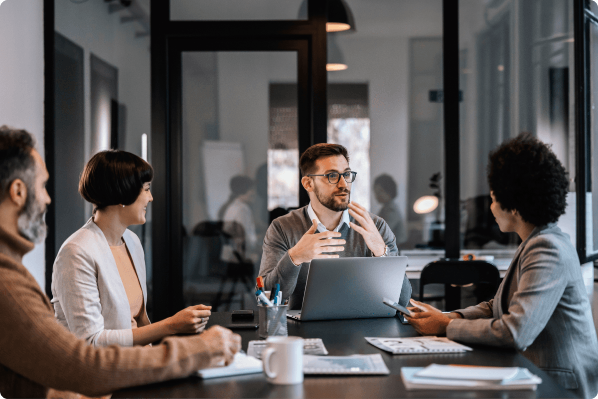 Four people sitting around a table in a conference room. The man at the head of the table has a laptop, and he is talking and gesturing with his hands. The others are looking at him.