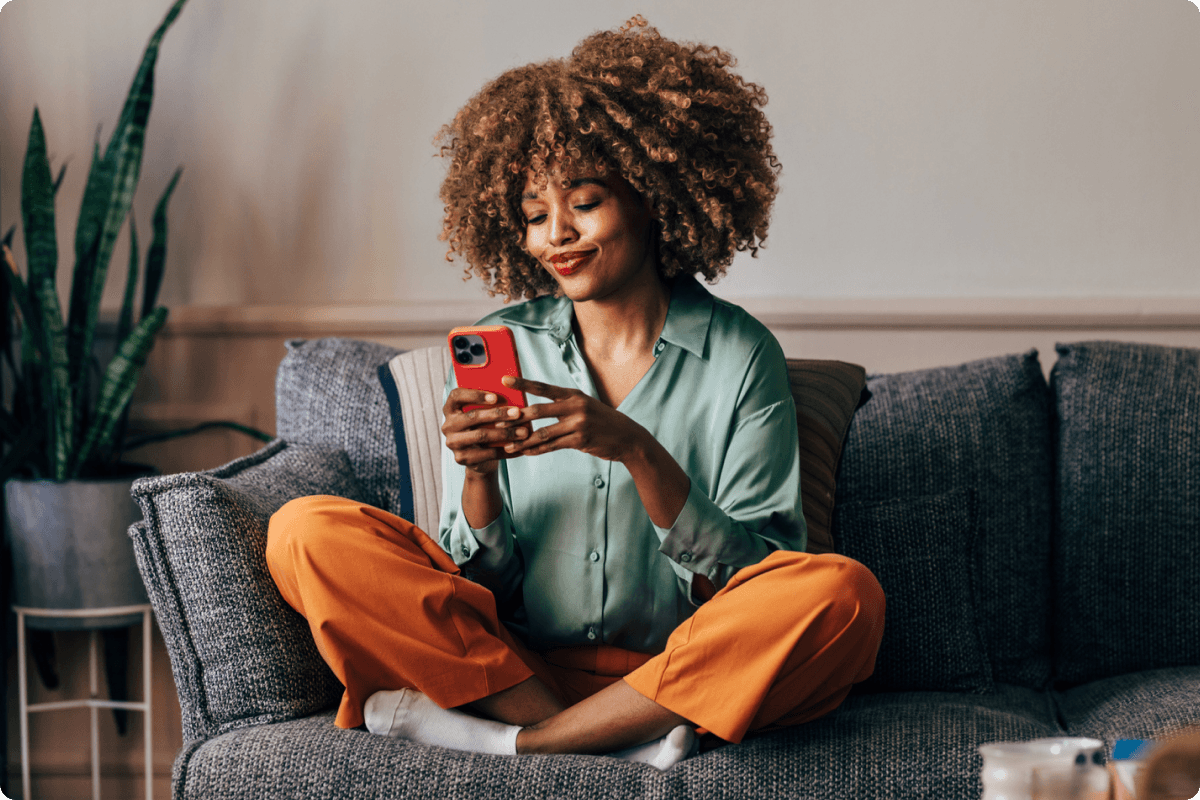 A woman sits comfortably on her couch, holding her cell phone and smiling while looking down at it.