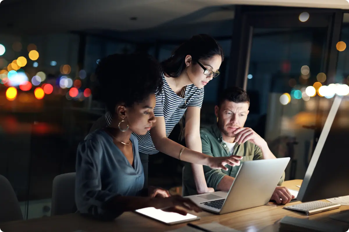 Photo of three employees working at a single laptop in a office at night.